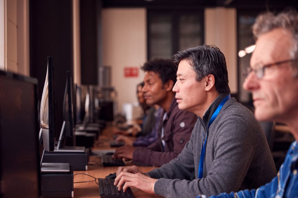 Group Of Mature Adult Students In Class Working At Computers In College Library